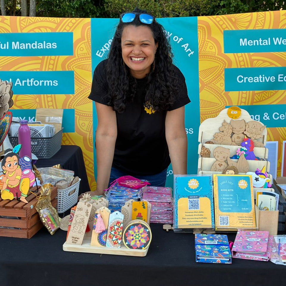 Woman standing behind a table with various items and a colorful banner in an outdoor setting.