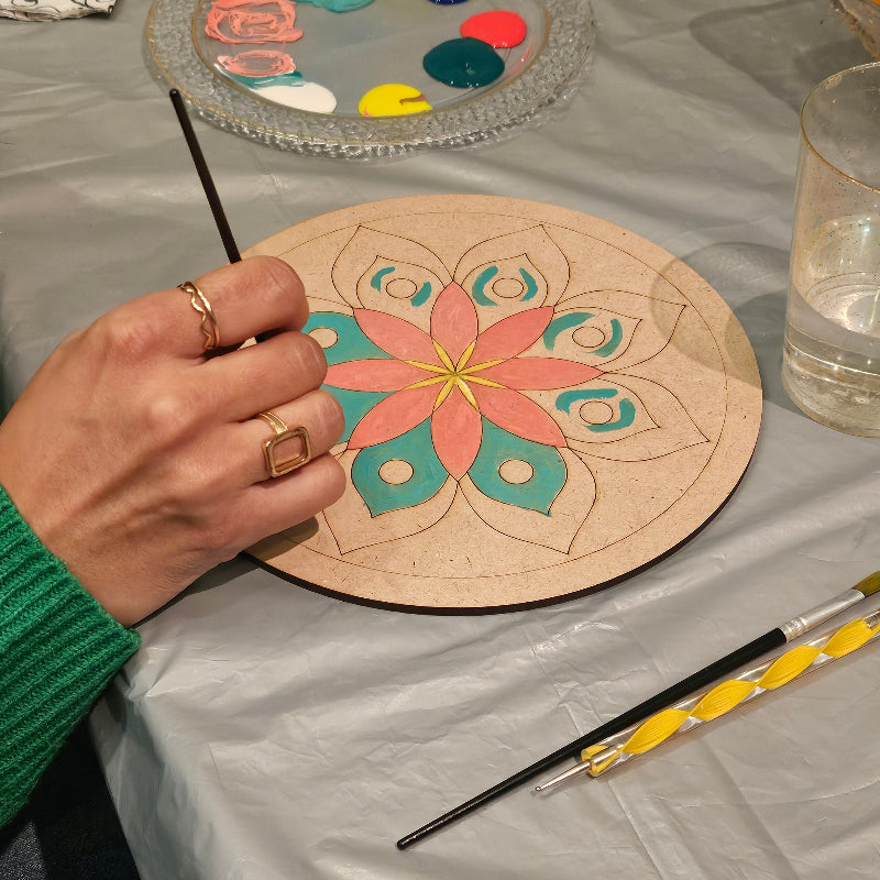 Person painting a mandala on a wooden board with a paintbrush, next to a colour palette and additional paint supplies.