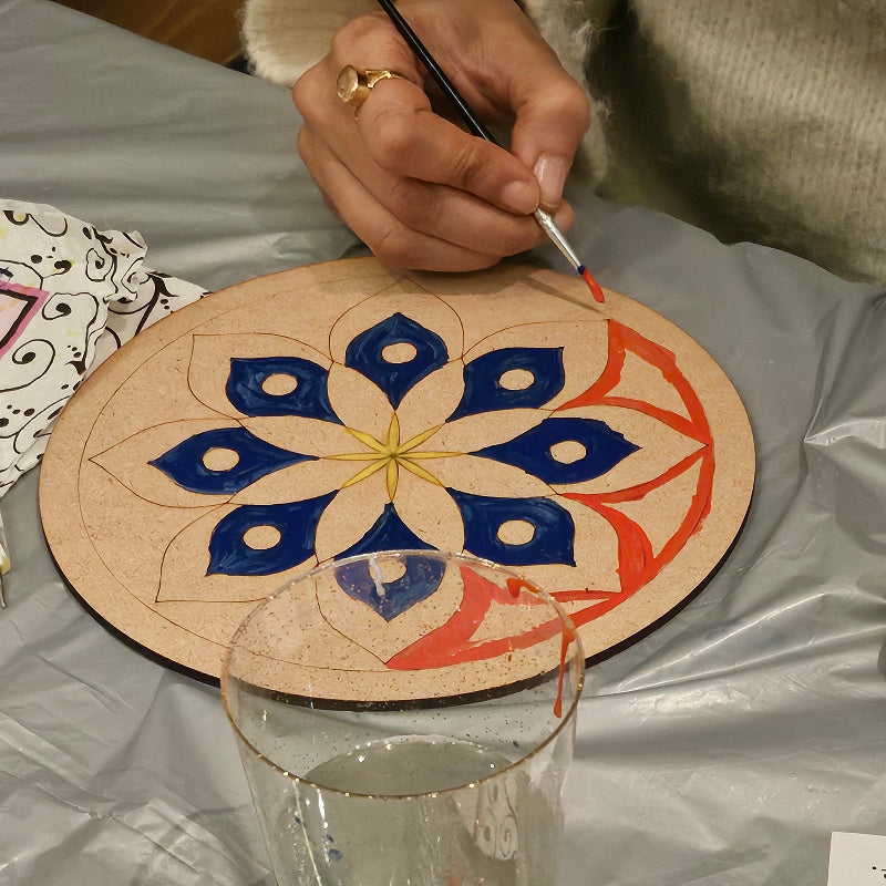 Person painting a decorative Mandala design on a wooden board with a glass container nearby.