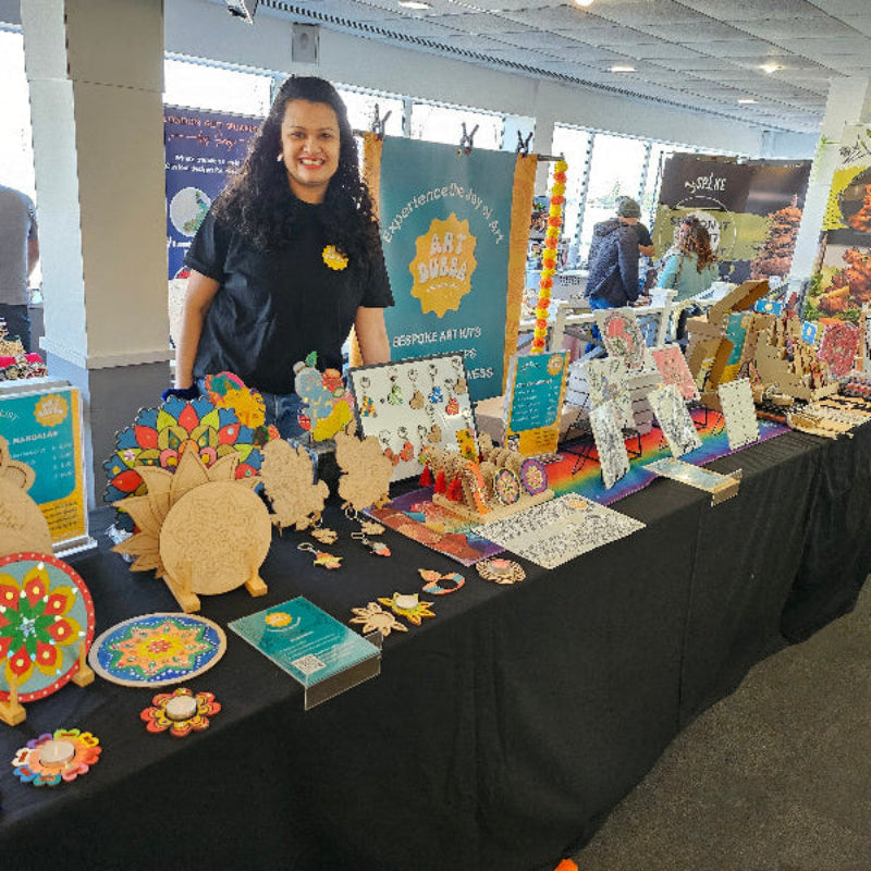 Founder of Art Dubba standing behind a table displaying various crafts and art items in an indoor setting.