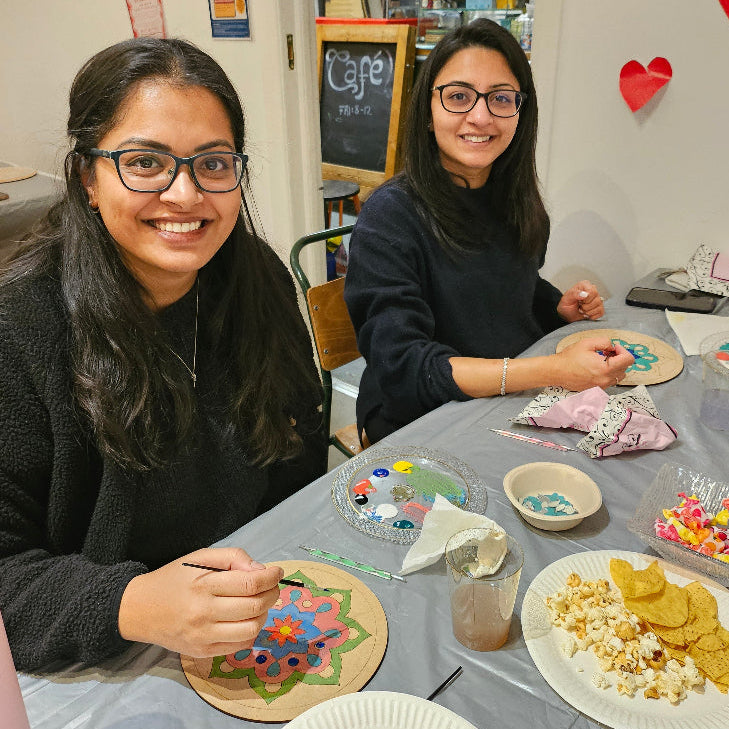 Two women at a table at a Mandala night workshop with Art supplies and snacks, smiling at the camera.