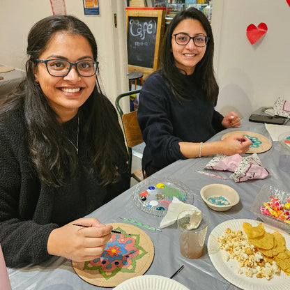 Two women at a table at a Mandala night workshop with Art supplies and snacks, smiling at the camera.