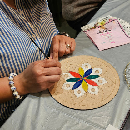 Person painting a round wooden mandala board with colourful design on a table with various items.