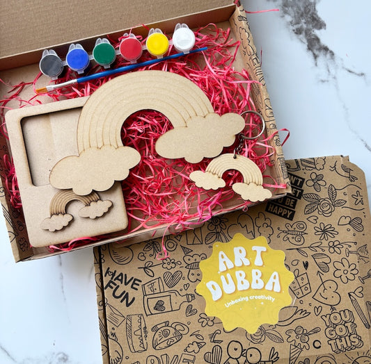 Wooden rainbow and cloud shapes with paint bottles and a box labeled 'Art Dubba' on a marble surface.
